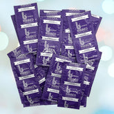 A pile of b Condoms Ribbed Condoms packets from bCondoms, featuring a bold and modern design crafted from premium natural rubber latex. The purple packaging displays white text that reads "Ribbed" along with other details against a soft, blurry gradient background adorned with circular light bokeh effects.